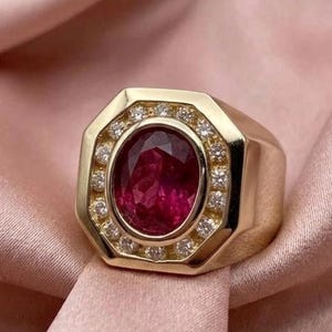 May include: A gold ring featuring a large, oval-cut deep red gemstone at the center, surrounded by a halo of small, round, clear white stones set within an octagonal frame. The band is thick and polished, showcasing a smooth, reflective gold finish.