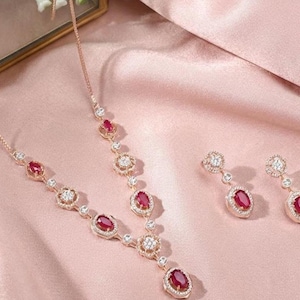 May include: A rose gold jewelry set consisting of a necklace and a pair of drop earrings. The necklace features alternating oval-cut red stones and round clear stones in a delicate chain. The earrings mirror this design with a single red stone.