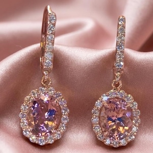 May include: A pair of rose gold drop earrings featuring oval-cut peach-coloured centre stones surrounded by a halo of small clear white crystals. The hinged lever-back closures are also accented with a row of small clear white crystals.