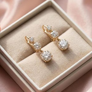 May include: Gold-coloured drop earrings, each with three clear, round gemstones. The earrings are presented in a white jewellery box with a beige interior, set against a soft pink background.