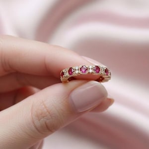 May include: A gold ring featuring a series of oval-cut, deep red gemstones, alternating with small, round, clear stones. The ring is set against a soft pink background, highlighting the jewelry's sparkle and design.