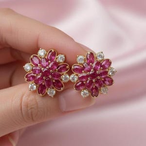 May include: A pair of gold-toned flower-shaped stud earrings. Each earring features a cluster of oval-cut, deep pink gemstones surrounded by smaller, round, clear gemstones. The earrings are held against a soft pink background.