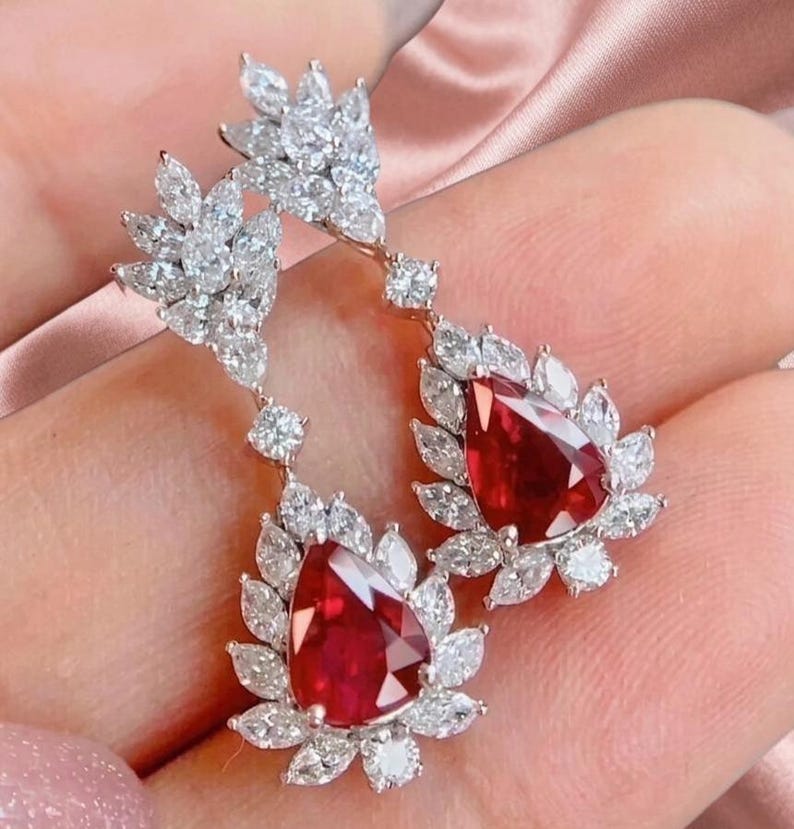 May include: A pair of drop earrings featuring two pear-shaped red gemstones, each surrounded by a cluster of marquise and round-cut white stones set in a metallic frame. The design includes a smaller upper section and a larger lower section.