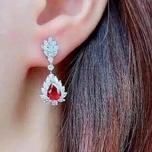 May include: A close-up of a drop earring with a large, red, teardrop-shaped gemstone, encircled by smaller, clear stones. The earring is attached to a smaller cluster of clear stones in a leaf design. The jewellery is set against a blurred background.