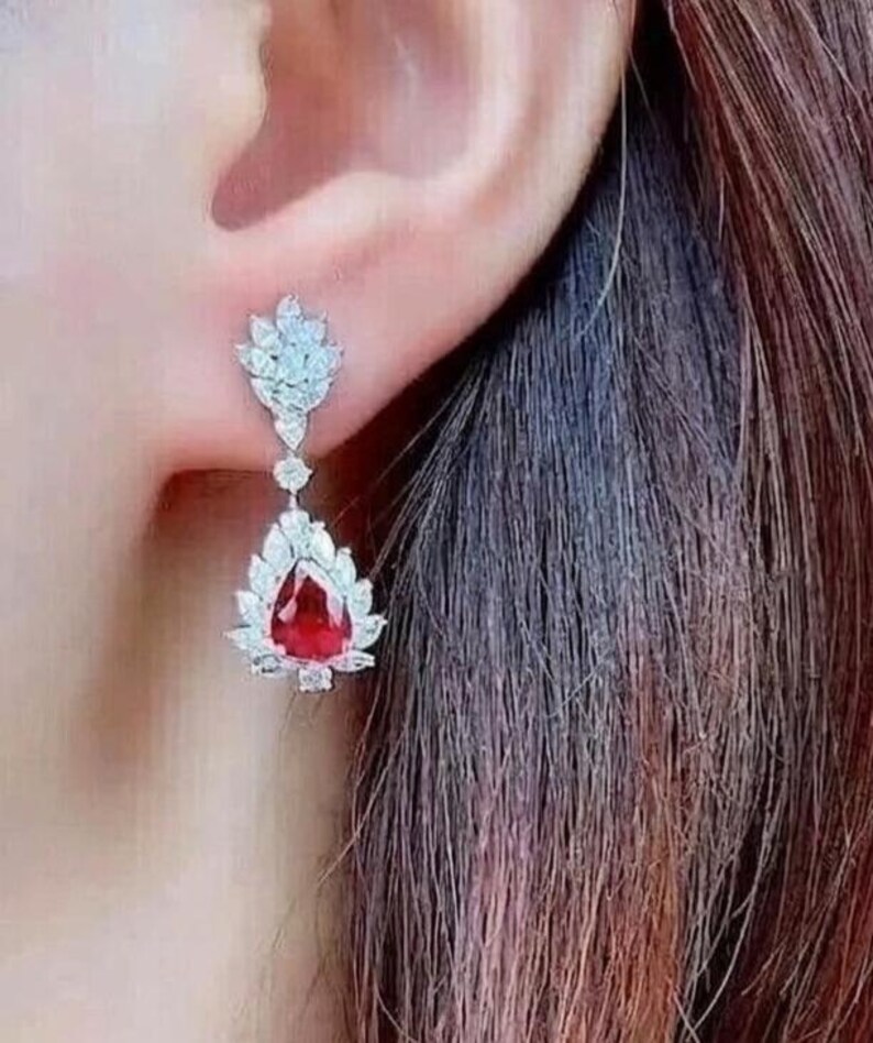May include: A close-up of a drop earring with a large, red, teardrop-shaped gemstone, encircled by smaller, clear stones. The earring is attached to a smaller cluster of clear stones in a leaf design. The jewellery is set against a blurred background.