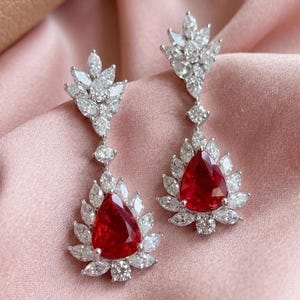 May include: A pair of drop earrings with a striking design. Each earring features a large, red, pear-cut gemstone, encircled by smaller white stones. The earrings have a floral motif and are set in a silver metal.