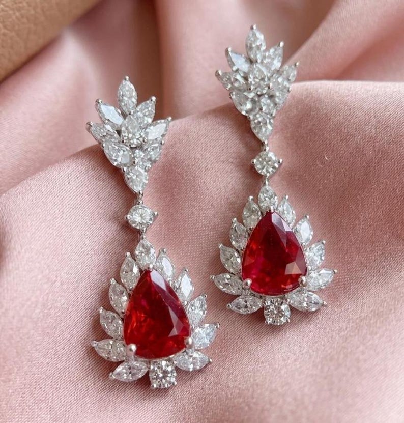 May include: A pair of drop earrings with a striking design. Each earring features a large, red, pear-cut gemstone, encircled by smaller white stones. The earrings have a floral motif and are set in a silver metal.