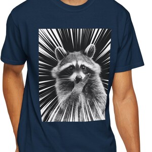 May include: Navy blue t-shirt featuring a black and white graphic of a raccoon with a radial burst design. The raccoon is in a close-up pose, with its paw near its mouth. The t-shirt is made of a soft material.
