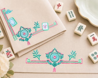 May include: A beige fabric napkin featuring intricate embroidery in shades of teal, pink, and green. The design includes geometric borders, stylised floral motifs, and bamboo-like sprigs. Several mahjong tiles with various symbols are scattered nearby.