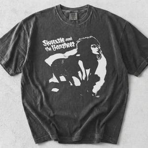 May include: Dark gray t-shirt featuring a black and white graphic of a woman and the text "Siouxsie and the Banshees". The shirt has a vintage, worn look.