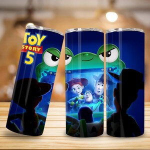 May include: A cylindrical tumbler featuring a dark blue background with the Toy Story 5 logo in yellow and red. The design displays a green frog-shaped screen showing animated characters including a cowgirl, a cowboy, and a space ranger.