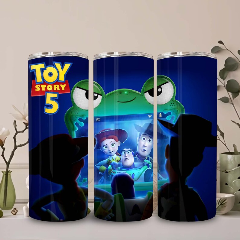 May include: A cylindrical tumbler featuring a dark blue background with the Toy Story 5 logo in yellow and red. The design displays a green frog-shaped device screen showing characters Jessie, Woody, and Buzz Lightyear in a dark setting.