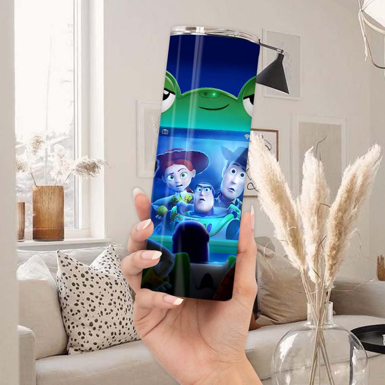 May include: A cylindrical metal tumbler featuring a vibrant graphic print of characters from the Toy Story film franchise, including Buzz Lightyear and Jessie, set against a blue background with a green alien character at the top.