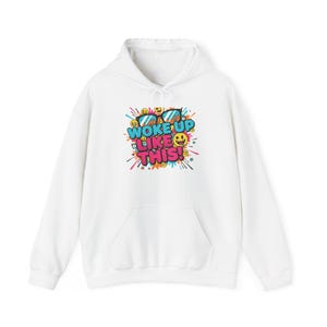 May include: White hoodie with a front pocket and a drawstring hood. The front displays a colourful graphic with sunglasses, emojis, and the text "WOKE UP LIKE THIS!". The design includes shades of blue, yellow, pink, and orange.