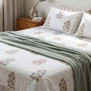 May include: White bed linen set with a floral pattern in pink and purple, and a sage green throw. The bedding includes two pillowcases. A wooden bedside table and a vase with a dried flower are also visible in the bedroom setting.