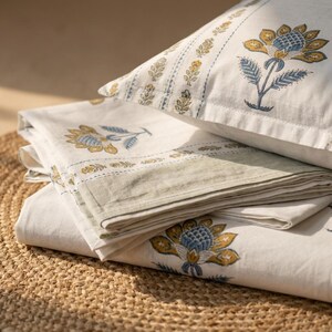 May include: A stack of white bedding with a floral pattern in yellow and blue. The bedding includes a pillowcase and folded sheets. The floral design is repeated across the fabric. The sheets have a light green border.