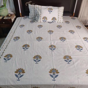 May include: White bedsheet set with a floral pattern in blue and gold. The set includes a fitted sheet and a pillowcase. The bedsheet has a repeating pattern of stylized flowers. The pillowcase has a similar design. The bed is against a dark brown headboard.