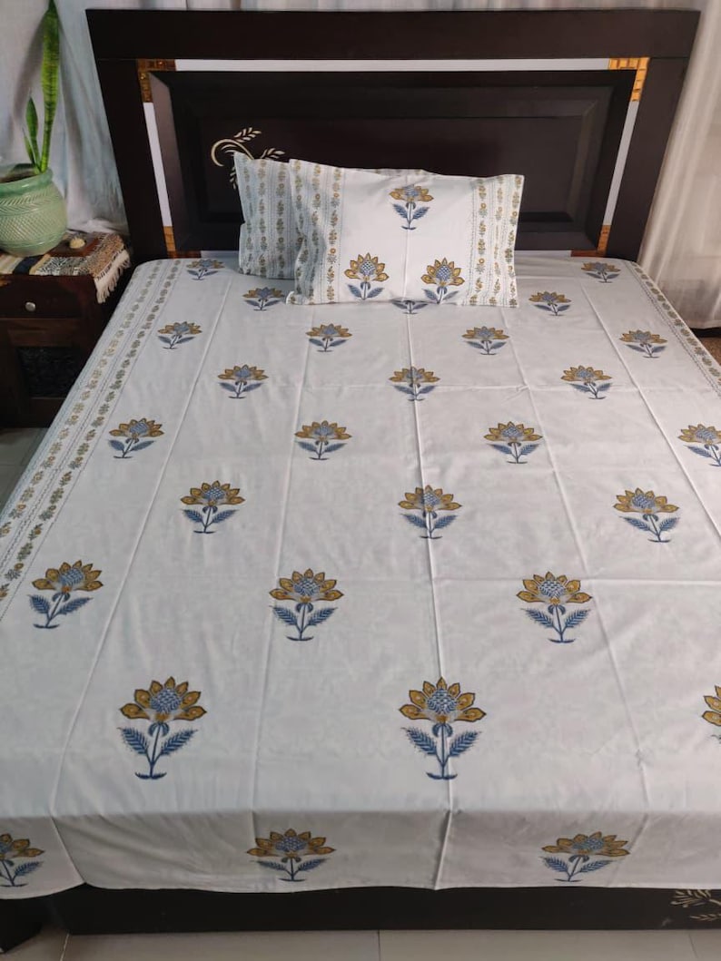 May include: White bedsheet set with a floral pattern in blue and gold. The set includes a fitted sheet and a pillowcase. The bedsheet has a repeating pattern of stylized flowers. The pillowcase has a similar design. The bed is against a dark brown headboard.