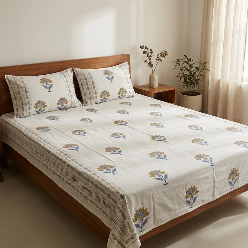 May include: A white floral patterned bed set featuring a duvet cover and two pillowcases. The design includes yellow and blue flowers with green stems. The bed set is on a wooden bed frame. The pillowcases and duvet cover have a decorative border.