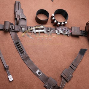 May include: A brown leather costume belt with multiple pouches, a holster, and a forearm guard. The belt includes two wrist cuffs and a shoulder strap. The accessories are arranged on a brown surface.