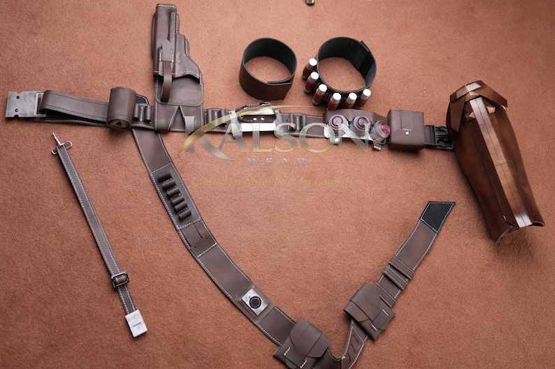 May include: A brown leather costume belt with multiple pouches, a holster, and a forearm guard. The belt includes two wrist cuffs and a shoulder strap. The accessories are arranged on a brown surface.