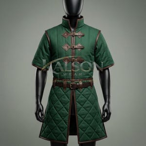 May include: A forest green quilted gambeson tunic featuring short sleeves, a high collar, and a front closure secured by three brown leather straps with metal buckles. A brown leather belt with a metal buckle is worn at the waist. Text reads ALSON WEAR.