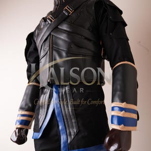 May include: A black faux leather vest with a high collar, diagonal straps, and metallic accents worn over a black shirt. The outfit includes black arm guards with blue and tan horizontal bands, and a black tunic with blue trim at the hem.