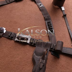May include: A brown leather utility belt with multiple pouches, holders, and a holster. The belt features white stitching and metal accents. The image also includes a matching shoulder strap and a cylindrical holder. The text "LALSON WEAR" is visible.