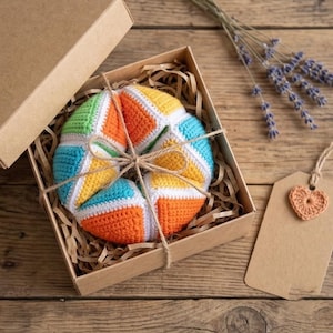 Puede incluir: Una colorida pelota de ganchillo en una caja de regalo de cartón. La pelota presenta segmentos verdes, naranjas, amarillos y azules, atados con hilo. Un pequeño corazón de ganchillo adorna una etiqueta en blanco. Lavanda seca y fondo de madera.