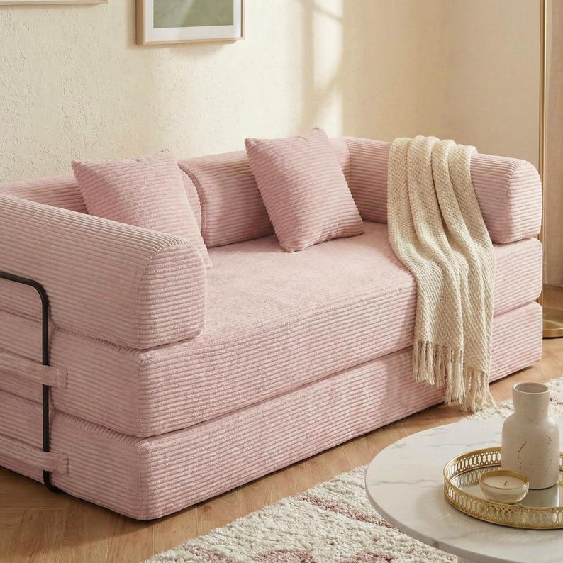 Puede incluir: Un sof&aacute; de pana rosa con cojines a juego y una manta de color crema. El sof&aacute; tiene un perfil bajo y un dise&ntilde;o minimalista, con una estructura de metal negro en el lateral. El sof&aacute; est&aacute; en un sal&oacute;n.