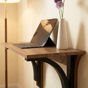 May include: A wooden wall-mounted desk with a black metal support. A black tablet with a keyboard is on the desk. A white vase with purple flowers sits on the desk. The desk is made of wood.