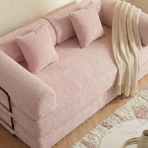 Puede incluir: Un sof&aacute; de pana rosa con cojines a juego y una manta color crema. El sof&aacute; tiene un perfil bajo y un dise&ntilde;o minimalista, adecuado para una sala de estar o zona de descanso. Los cojines son cuadrados y la manta tiene flecos.