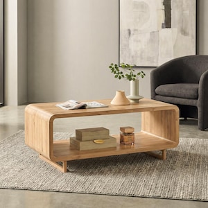 May include: A light-colored wooden coffee table with a lower shelf. The table has rounded edges and a rectangular shape. A magazine, vases, and decorative boxes are on the table. The table is in a living room setting.