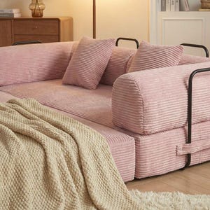 Puede incluir: Un sof&aacute; modular de pana rosa con reposabrazos de metal negro y varias almohadas a juego. Una manta de punto color crema est&aacute; sobre el sof&aacute;. El sof&aacute; est&aacute; en una habitaci&oacute;n bien iluminada con muebles de madera.