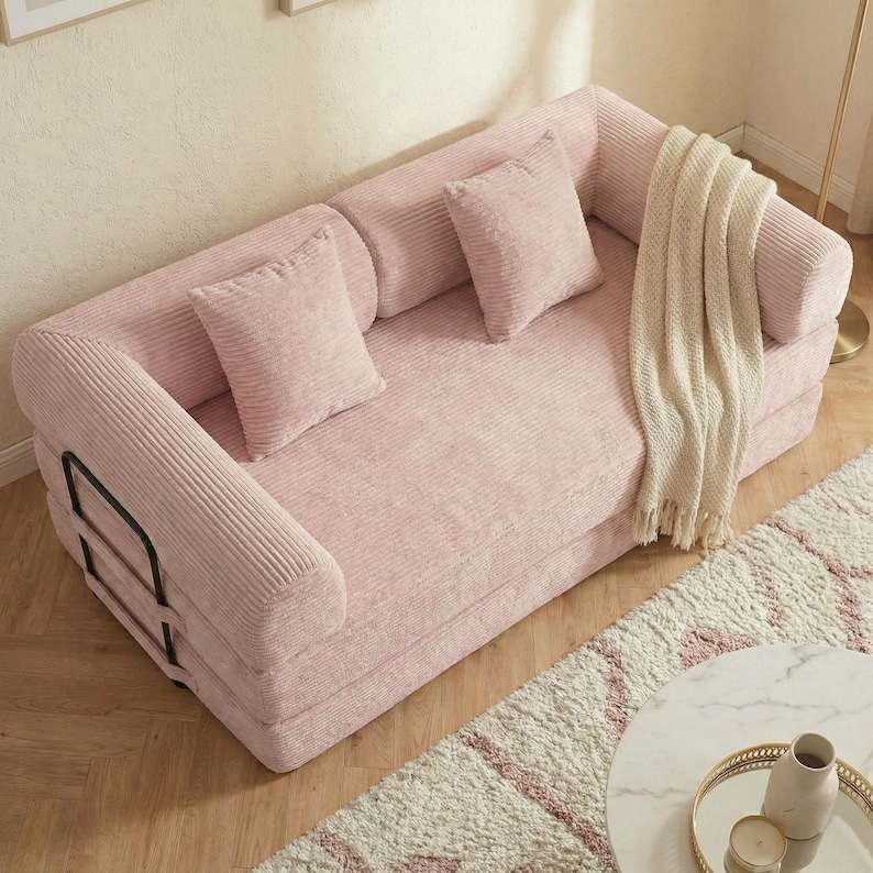 Puede incluir: Un sof&aacute; de pana rosa con dos cojines cuadrados a juego y una manta crema. El sof&aacute; tiene reposabrazos redondeados y una estructura met&aacute;lica negra. El sof&aacute; est&aacute; sobre una alfombra estampada.