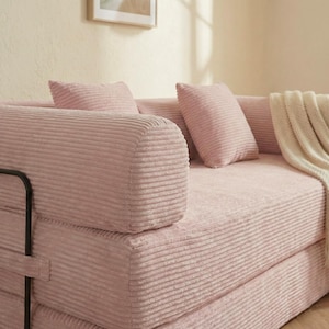Puede incluir: Un sof&aacute; de pana rosa con dos cojines a juego y una manta doblada. El sof&aacute; tiene un reposabrazos redondeado y una estructura de metal negro. La tela de pana tiene una textura acanalada. Los cojines son rectangulares y combinan con el color.