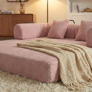 Puede incluir: Un sof&aacute; seccional de pana rosa con cojines a juego y una manta de punto beige. El sof&aacute; de perfil bajo est&aacute; sobre una alfombra de pelo largo blanca. La habitaci&oacute;n est&aacute; iluminada por una l&aacute;mpara c&aacute;lida.