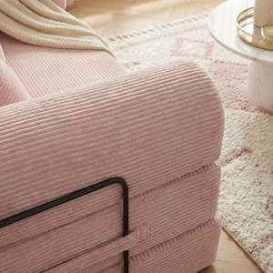 Puede incluir: Primer plano de un brazo de sof&aacute; de pana rosa con una estructura met&aacute;lica negra. El sof&aacute; tiene una textura acanalada y forma parte de un conjunto de asientos m&aacute;s grande. Una manta color crema y una alfombra estampada son visibles en el fondo.