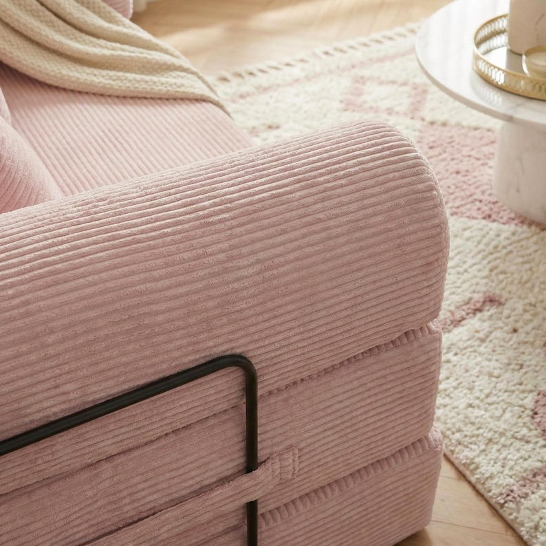 Puede incluir: Primer plano de un brazo de sof&aacute; de pana rosa con una estructura met&aacute;lica negra. El sof&aacute; tiene una textura acanalada y forma parte de un conjunto de asientos m&aacute;s grande. Una manta color crema y una alfombra estampada son visibles en el fondo.