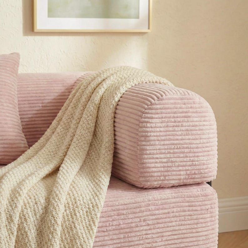 Puede incluir: Primer plano de un sof&aacute; de pana rosa con una manta de punto color crema. El sof&aacute; tiene un reposabrazos redondeado y un coj&iacute;n a juego. El fondo es una pared beige claro con un cuadro enmarcado.