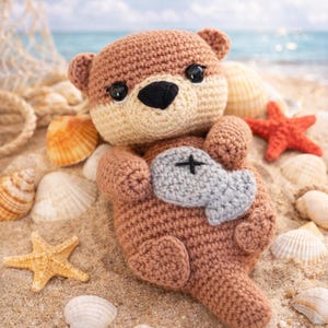 May include: A crocheted brown otter plush toy with a light beige face and a gray fish. The otter is lying on its back on a sandy beach, surrounded by seashells and starfish. The background shows the ocean and sky.