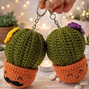 Puede incluir: Dos llaveros de cactus hechos a mano con ganchillo. Los cactus son verdes con detalles amarillos y morados, sentados en macetas naranjas con caras sonrientes y bigotes negros. Los llaveros están sujetos por un llavero plateado.