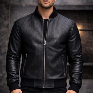 Op de afbeelding: Zwarte leren bomberjack met een getextureerd, reptielachtig patroon. De jas heeft een ritssluiting, twee zijzakken en geribde manchetten en zoom. Het model draagt een zwarte jeans en een zwart shirt.