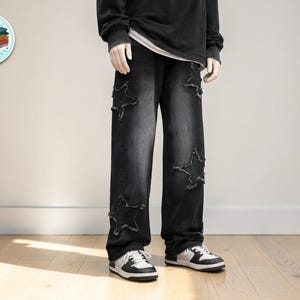 May include: Black denim jeans with star cutouts on the knees. The jeans have a relaxed fit and are paired with white and black sneakers. The model is wearing a black long-sleeve shirt.