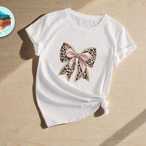 Puede incluir: Camiseta blanca con un diseño de lazo grande. El lazo tiene un estampado de leopardo con detalles de cinta rosa y ribete dorado. La camiseta se muestra sobre una superficie de madera clara.