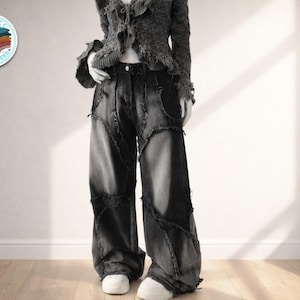 Può includere: Una persona indossa un maglione grigio scuro con frange e jeans in denim a gamba larga con patchwork. I jeans hanno un aspetto invecchiato con cuciture e toppe visibili. Le scarpe da ginnastica bianche completano l'outfit. Un logo è visibile.