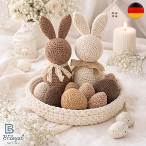 Puede incluir: Dos conejos de Pascua de ganchillo, uno marrón y otro crema, están en una cesta tejida con huevos de ganchillo en varios colores neutros. El conejo marrón tiene una cinta de encaje, y el conejo crema tiene una cinta de arpillera. Flores blancas rodean la cesta.