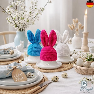 Könnte beinhalten: Eine festliche Ostertafel mit drei gehäkelten Hasen-Eierwärmern in Blau, Pink und Weiß. Die Hasen sitzen auf weißen Eierbechern, umgeben von Blumen, Kerzen und dekorierten Eiern. Ein Etikett trägt die Aufschrift "Glad Påsk".