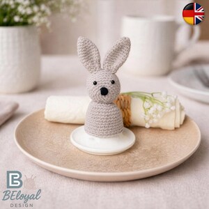 Könnte beinhalten: Eine gehäkelte, graue Hasenfigur mit schwarzen Augen und einer schwarzen Nase sitzt auf einem weißen Keramiksockel. Der Hase steht auf einem beigefarbenen Teller mit einer gerollten weißen Serviette und Grünzeug.