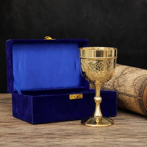 May include: A polished gold-tone metal chalice featuring intricate floral engravings on the bowl and a beaded rim, presented alongside a rectangular blue velvet-lined storage box with a gold-colored metal latch and a gold-colored hinge.